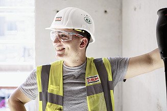 Karriere Ausbildung Industriemechaniker:in ZÜBLIN Timber Bild eines jungen Mannes in Arbeitskleidung. Er lehnt an einer Wand und blickt zur Seite.