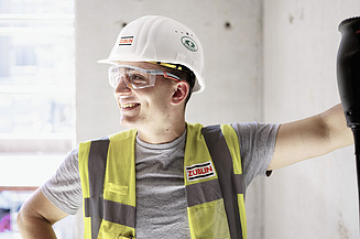 Picture of a young man in work clothes. He is leaning against a wall and looking to the side.