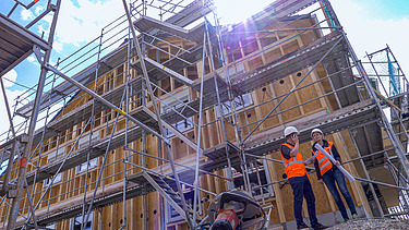Foto von zwei Personen auf einer Baustelle. Im Hintergrund ist ein Holzgebäude.