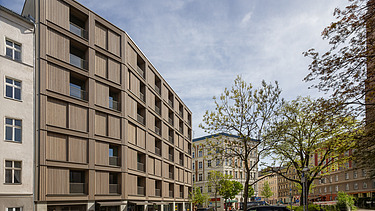 Foto der mehrgeschossigen Wohnanlage in der Stendaler Straße Berlin. Man sieht ein hohes Gebäude mit Holzfassade.