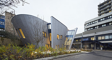 Foto eines Gebäudes mit Holzfassade und schrägen Wänden. Man sieht ein weiteres großes Gebäude auf dem Bild.