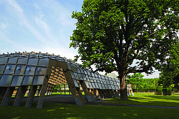 Foto des Serpentine Pavillons in London. Man sieht eine offene, begehbare Holzkonstruktion auf einer grünen Wiese mit einem Baum.