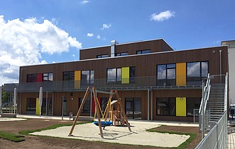 Photo of a wooden school from the outside. You can see the wooden building and the garden area with a swing.