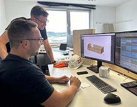 Photo of two men at a computer workstation. One is sitting, the other is standing.