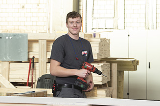 Foto eines jungen Auszubildenden Zimmerers, der in der Produktion steht und eine Bohrmaschine hält.