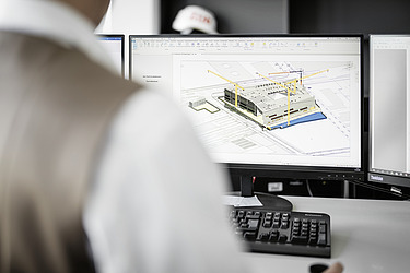 Photo of a computer workstation with a sketch of a building on the screen. A person sitting in front of it can be seen from behind.