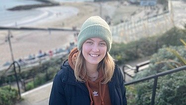 Foto einer jungen Frau. Im Hintergrund ist der Strand und das Meer zu sehen.