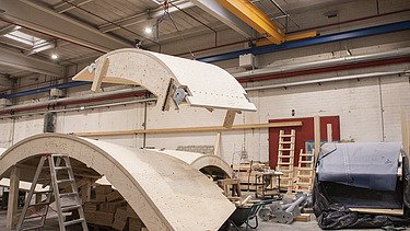 Photo of a production hall with two curved wooden shells, one hanging from the crane