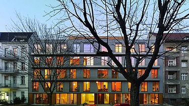 Foto eines mehrgeschossigen Gebäudes in dämmeriger Lichtstimmung. Zuvor stehen blattlose Bäume.