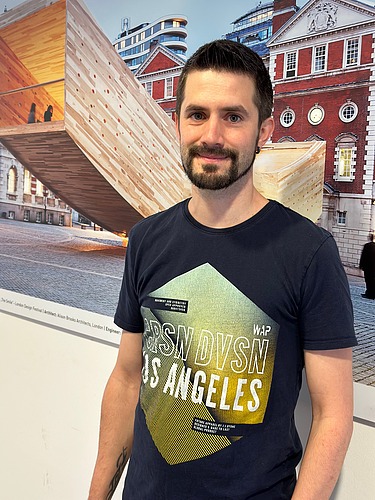 Foto eines Mannes mit dunklen kurzen Haaren und kurzem Bart. Er trägt ein T-Shirt mit Aufdruck. Im Hintergrund sieht man ein Bild mit einer geschwungenen Holzskulptur.