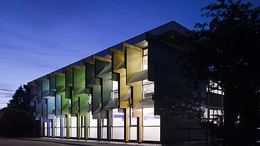 Nachtaufnahme eines Schulgebäudes mit bunten Fassadenelementen aus Furnierschichtholz.