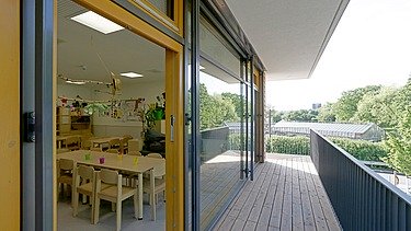 Innenaufnahme der Kita in Frankfurt. Man sieht eine Terrasse und durch ein geöffnetes Fenster einen Aufenthaltsraum. 