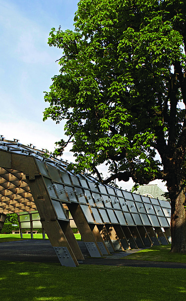 Foto des Serpentine Pavillons in London. Man sieht eine offene, begehbare Holzkonstruktion auf einer grünen Wiese mit einem Baum.