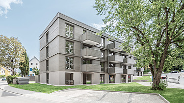 Foto einer Außenaufnahme eines Holzgebäudes mit Holzfassade und einem großen Laubbaum davor.