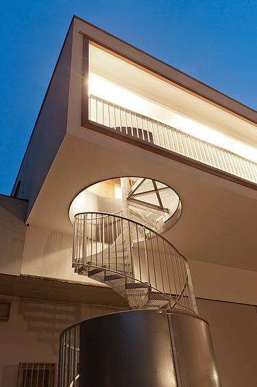 Night shot of a section of a building with external spiral staircase. Wooden extension.