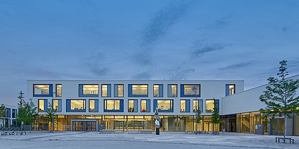 Außenaufnahme des Gymnasiums München-Nord. Man sieht ein großes, weißes Gebäude mit Flachdach in dämmriger Abendstimmung.