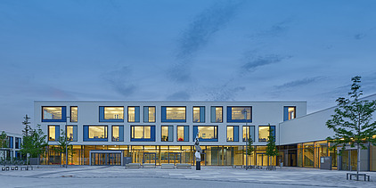Exterior shot of the Munich North Grammar School. You can see a large, white building with a flat roof in a dusky evening atmosphere.