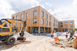 Photo of Witten University from the outside. You can see a large wooden building, an excavator and people.