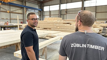 Foto von zwei jungen Männern, die in der Produktion eines Holzbaubetriebs stehen.