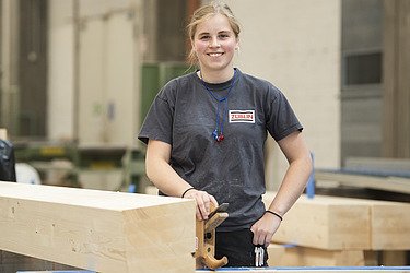 Foto einer jungen Zimmerer-Kollegin, die in der Holzproduktionshalle steht und ein Holzelement schleift.