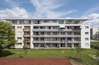 Photo of a multi-storey building with garden and hard court in front. Wooden extension.