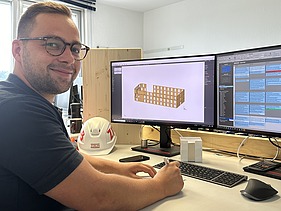Photo of a young man sitting at a computer workstation and smiling at the camera.