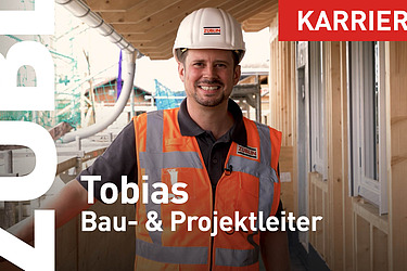 Foto von einem Mann mit Bauhelm und Arbeitskleidung. Ein Holzgebäude ist zu sehen.