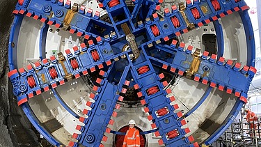 Foto von einer jungen Frau in oranger Arbeitskleidung vor einer rießengroßen Tunnelbohrmaschine in blau-rot.
