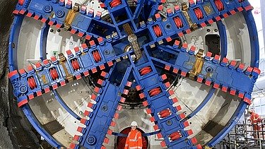 Foto von einer jungen Frau in oranger Arbeitskleidung vor einer rießengroßen Tunnelbohrmaschine in blau-rot.