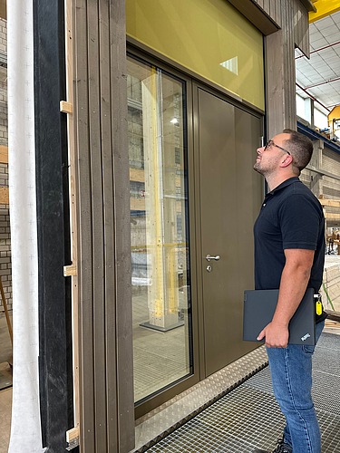 Foto von einem jungen Mann der vor einem Holzelement in der Produktion seht und es betrachtet.