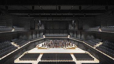 Photo of an interior of the interim venue Gasteig in Munich. You can see black wooden acoustic elements on the walls and ceiling and the orchestra in the middle.
