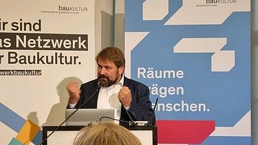 Photo of a man at the lectern. Roll-ups can be seen in the background.