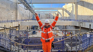 Foto von einer Frau in orangener Arbeitskleidung und weißem Helm auf dem Kopf. Siie streckt beide Hände nach oben und lächelt. Im Hintergrund sieht man eine Baustelle aus Beton.