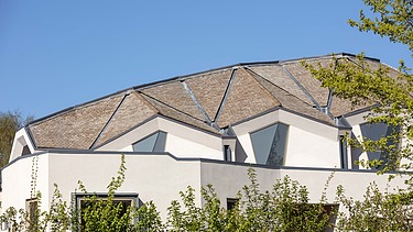 Foto eines Gebäudes mit weißer Fassade und Dach mit Gauben. Blauer Himmel und Sträucher sind zu sehen.