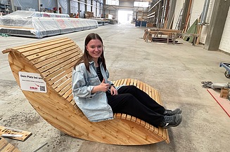 Foto einer jungen Frau in der Werkshalle von ZÜBLIN Timber. Sie sitzt auf einer Holzliege.