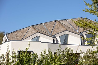 Foto eines Gebäudes mit weißer Fassade und Dach mit Gauben. Blauer Himmel und Sträucher sind zu sehen.