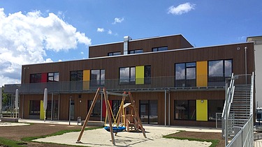 Photo of a wooden school from the outside. You can see the wooden building and the garden area with a swing.