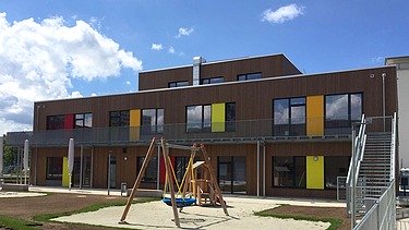 Photo of a wooden school from the outside. You can see the wooden building and the garden area with a swing.