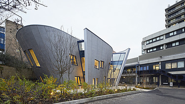 Foto eines Gebäudes mit Holzfassade und schrägen Wänden. Man sieht ein weiteres großes Gebäude auf dem Bild.