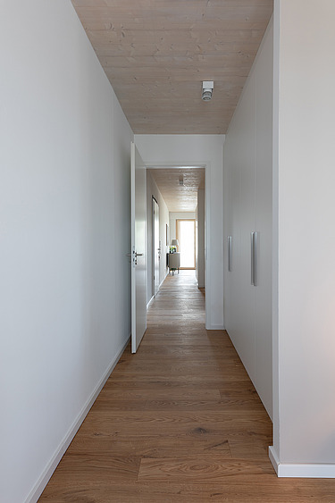Foto eines leeren, weißen Flures im Holzhochhaus CARL. Man sieht Holzoberflächen auf dem Boden und an der Decke