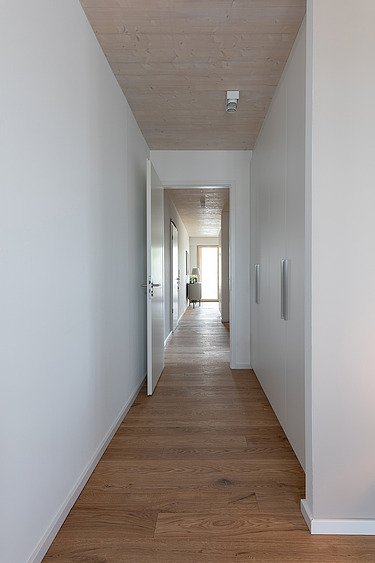 Foto eines leeren, weißen Flures im Holzhochhaus CARL. Man sieht Holzoberflächen auf dem Boden und an der Decke