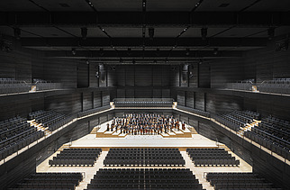 Photo of an interior of the interim venue Gasteig in Munich. You can see black wooden acoustic elements on the walls and ceiling and the orchestra in the middle.