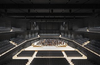 Photo of an interior of the interim venue Gasteig in Munich. You can see black wooden acoustic elements on the walls and ceiling and the orchestra in the middle.
