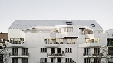 Foto einer Außenaufnahme eines mehrgeschossigen Gebäudes mit Aufstockung aus Holz. Das Gebäude hat eine weiße Fassade.