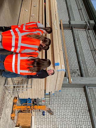 Foto von mehreren jungen Frauen und einem Mann, die in der Produktionshalle von ZÜBLIN Timber an einem LENO-Holzstapel stehen.