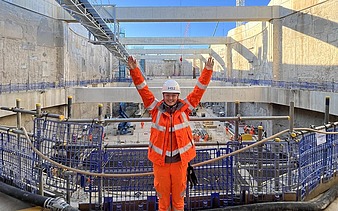 Foto von einer Frau in orangener Arbeitskleidung und weißem Helm auf dem Kopf. Siie streckt beide Hände nach oben und lächelt. Im Hintergrund sieht man eine Baustelle aus Beton.