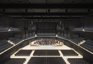 Foto der Interimsspielstätte Gasteig in München. Zu sehen ist der bestuhlte Bühnenraum mit Orchester.