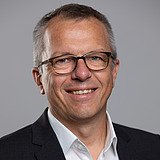 Photo of a smiling man with glasses, black jacket and white shirt. Heinz Thurik.