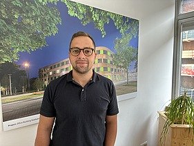 Foto von einem jungen Mann. Im Hintergrund sieht man ein Bild mit einem Holzgebäude darauf.