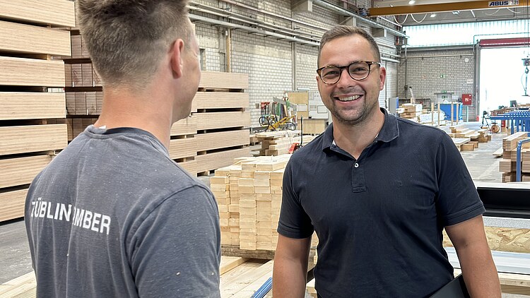 Foto von zwei jungen Männern in einer Holzproduktionshalle. Einer lächelt in die Kamera, der Andere zeigt seinen Rücken.